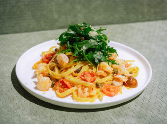 Plate of fettuccine pasta topped with shrimp, tomatoes, arugula, and parmesan cheese on a white plate.